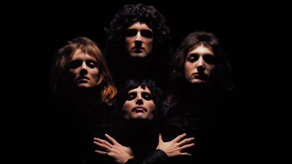 Os quatro integrantes da banda Queen em sombras, com luz focada em seus rostos, na clássica pose de abertura do clipe de Bohemian Rhapsody.